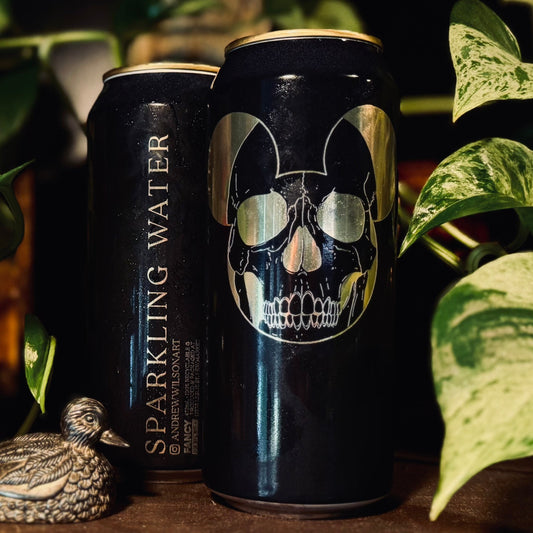 Two dark beverage cans on a wooden surface surrounded by green houseplants. The left can reads "SPARKLING WATER" vertically, and the right can features a large metallic silver skull graphic with cat-like ears on a black background.